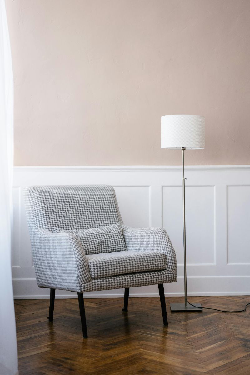 A patterned armchair next to a floor lamp against a light beige wall with white wainscoting and dark wood flooring.