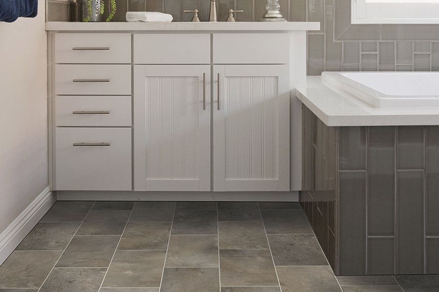 Bathroom vanity with white cabinets, gray tile floor, and a white countertop beside a tiled tub wall