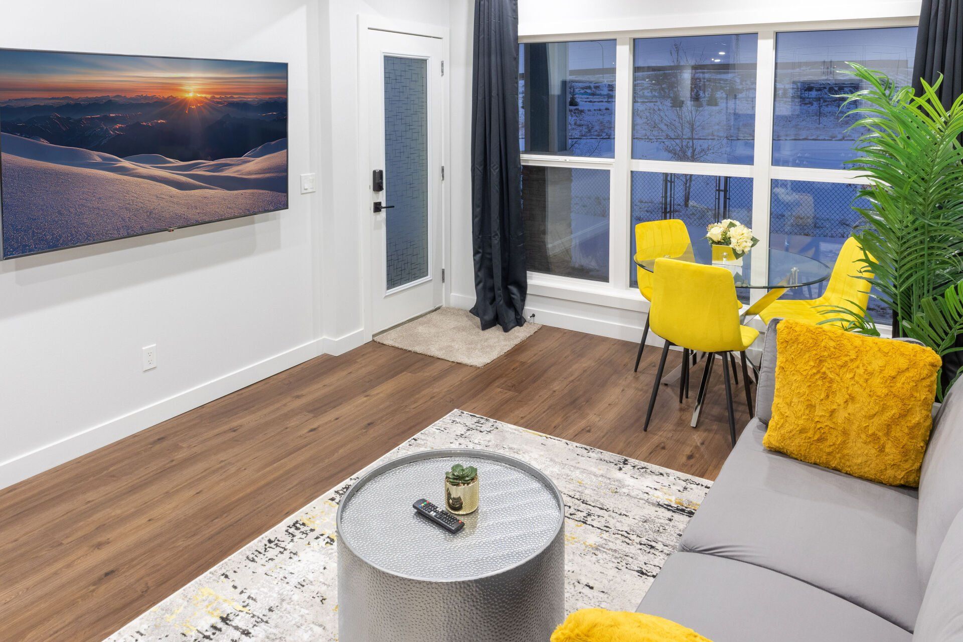 picture shows 3 seat couch, a glass round dinning table with 3 yellow chairs, an area carpet and a TV hung on a wall in a Calgary basement development