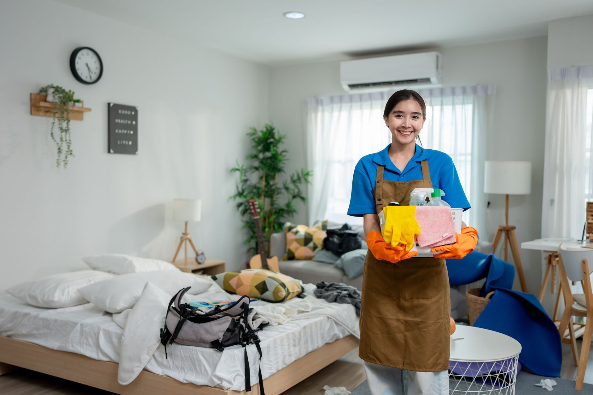 Smiling woman in apron holds cleaning supplies in a messy room.