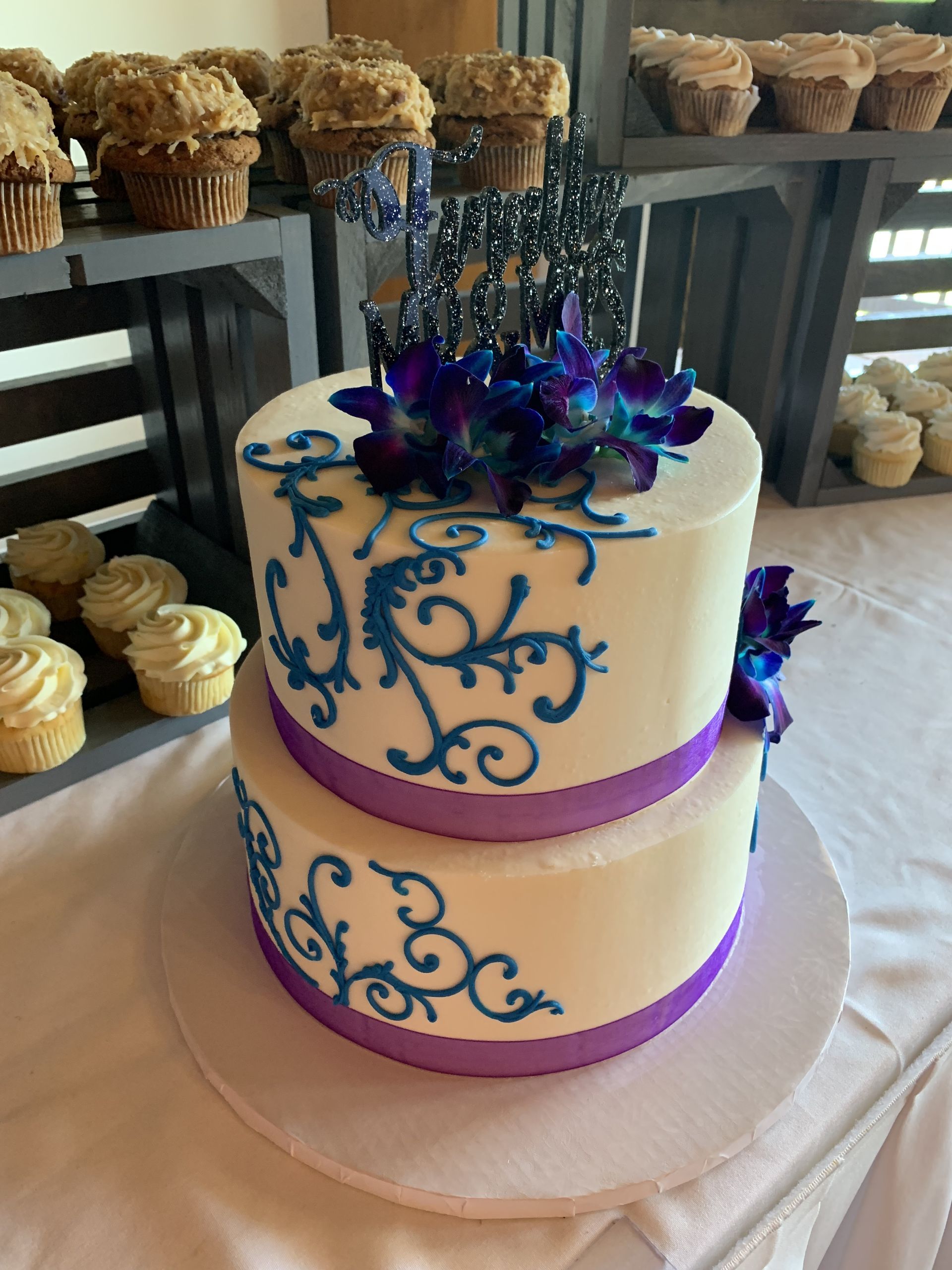 a wedding cake with purple and blue flowers is on a table with cupcakes .