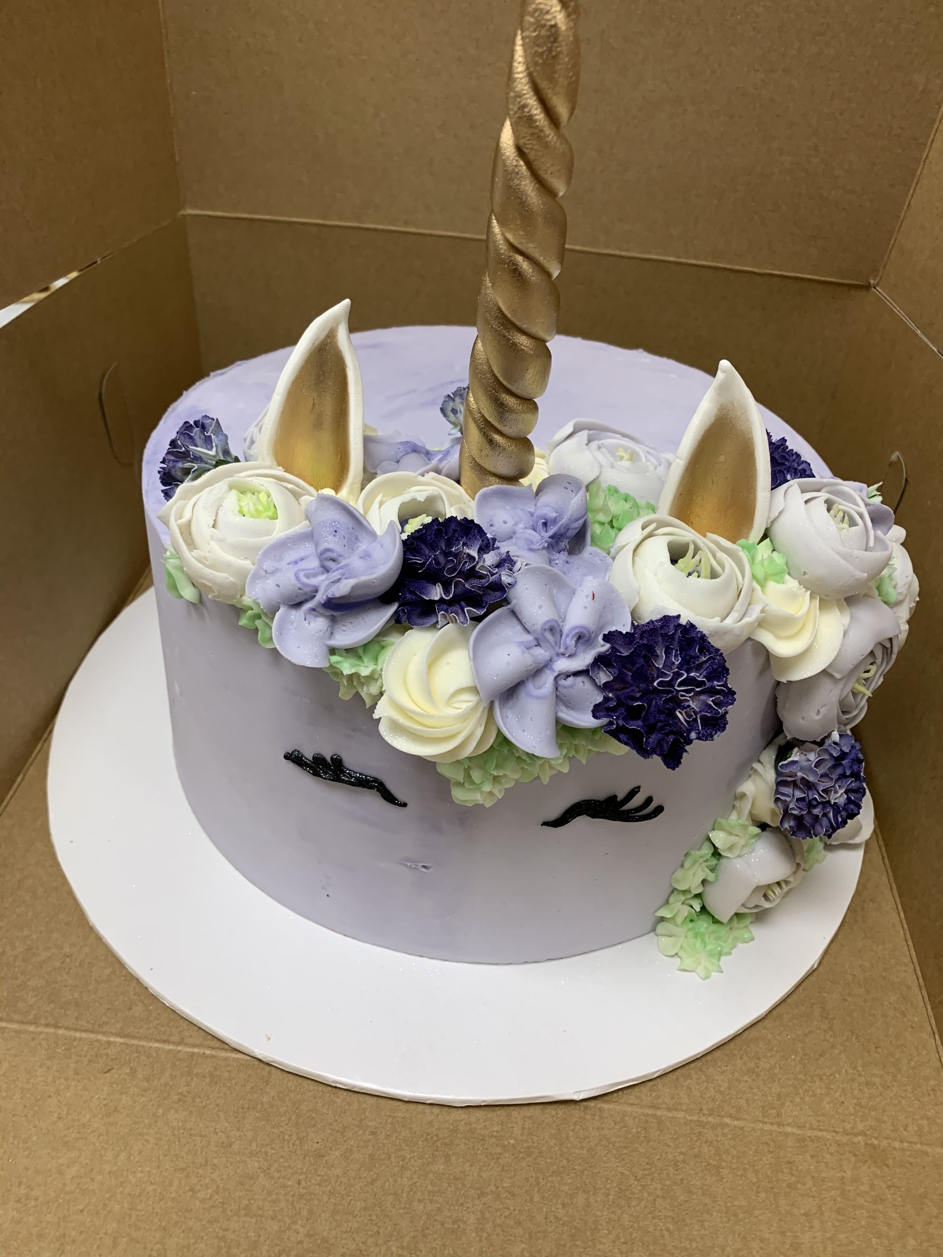 a unicorn cake with purple flowers and a gold horn is in a box .