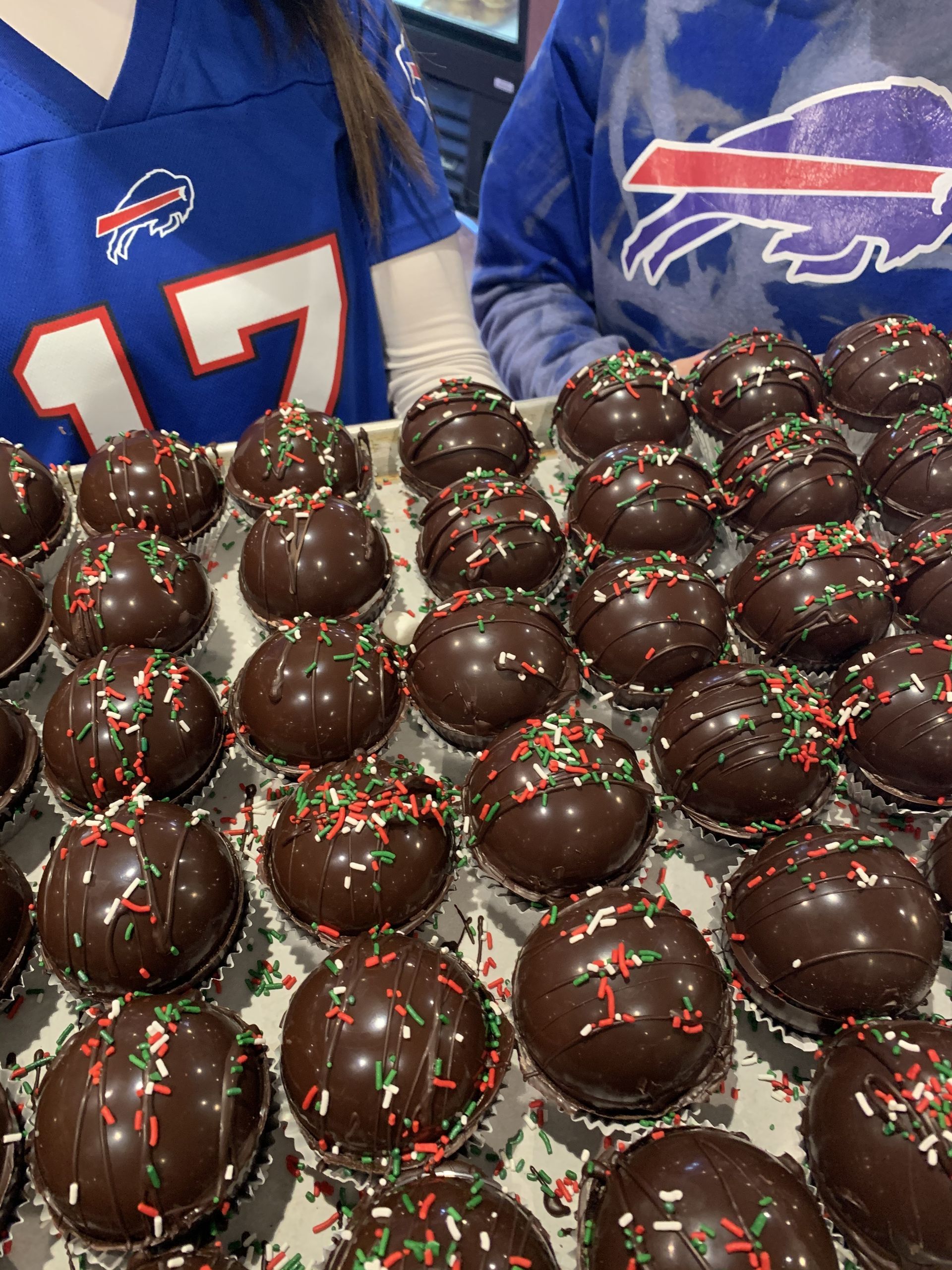 a person wearing a buffalo bills jersey is standing next to a tray of chocolate covered cupcakes .