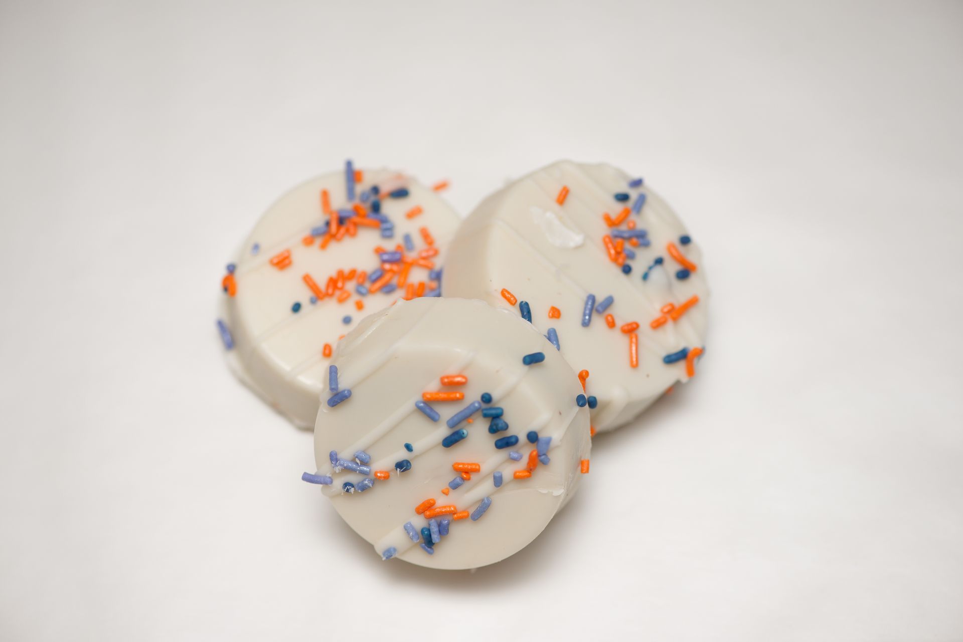 three white chocolate covered cookies with blue and orange sprinkles on a white surface .