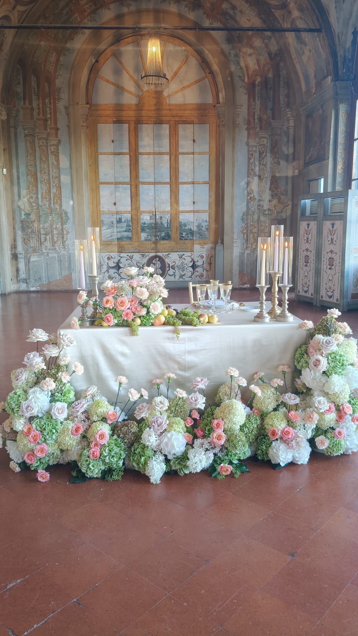 Tre grandi e lussureggianti composizioni floreali bianche in alti vasi di vetro, disposte su un tavolo apparecchiato con una tovaglia bianca in una location all'aperto.
