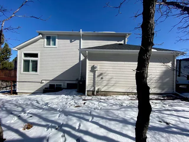 A white house with a garage is surrounded by snow and trees.