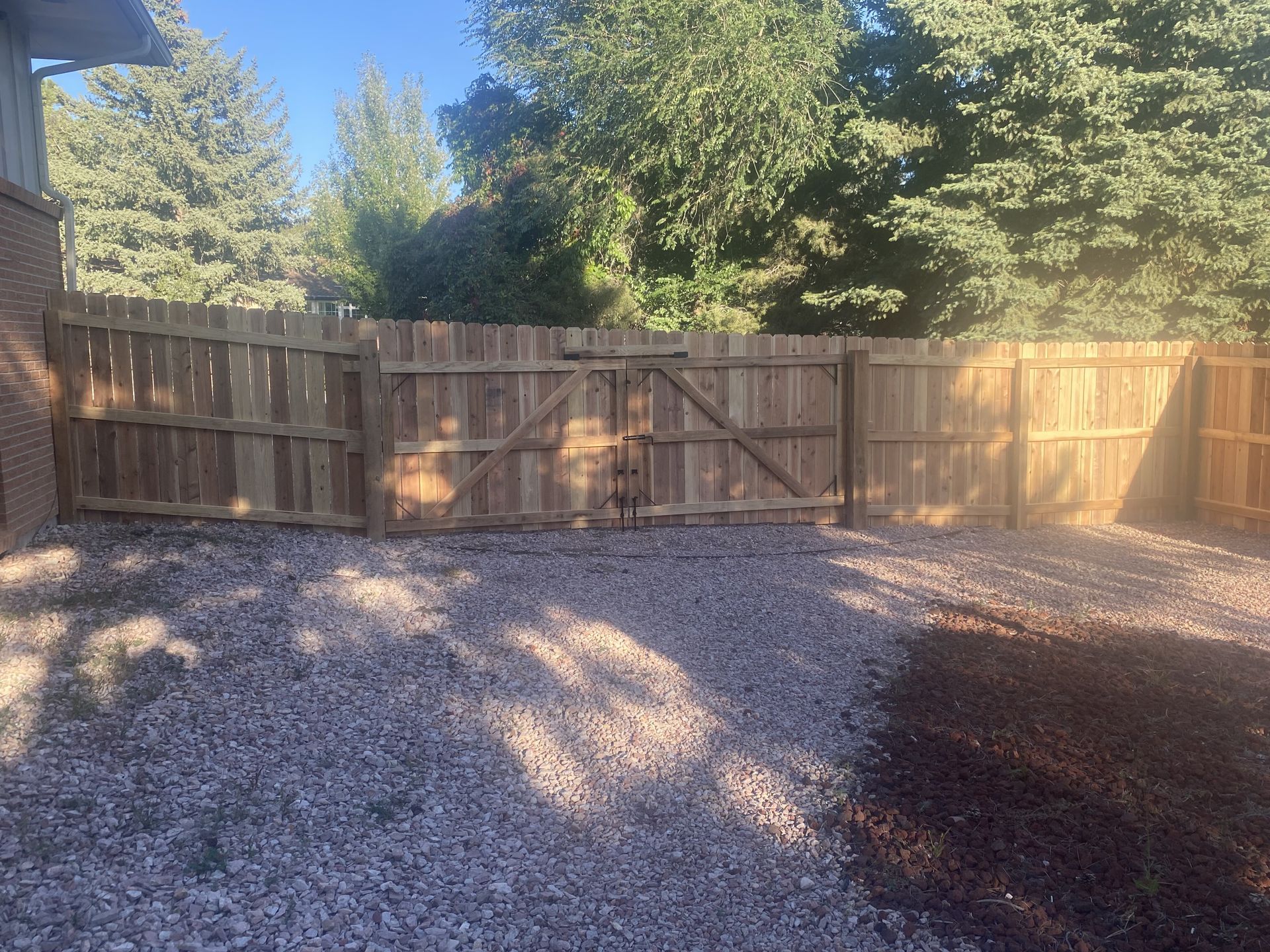 A wooden fence is surrounded by gravel and trees in a backyard.