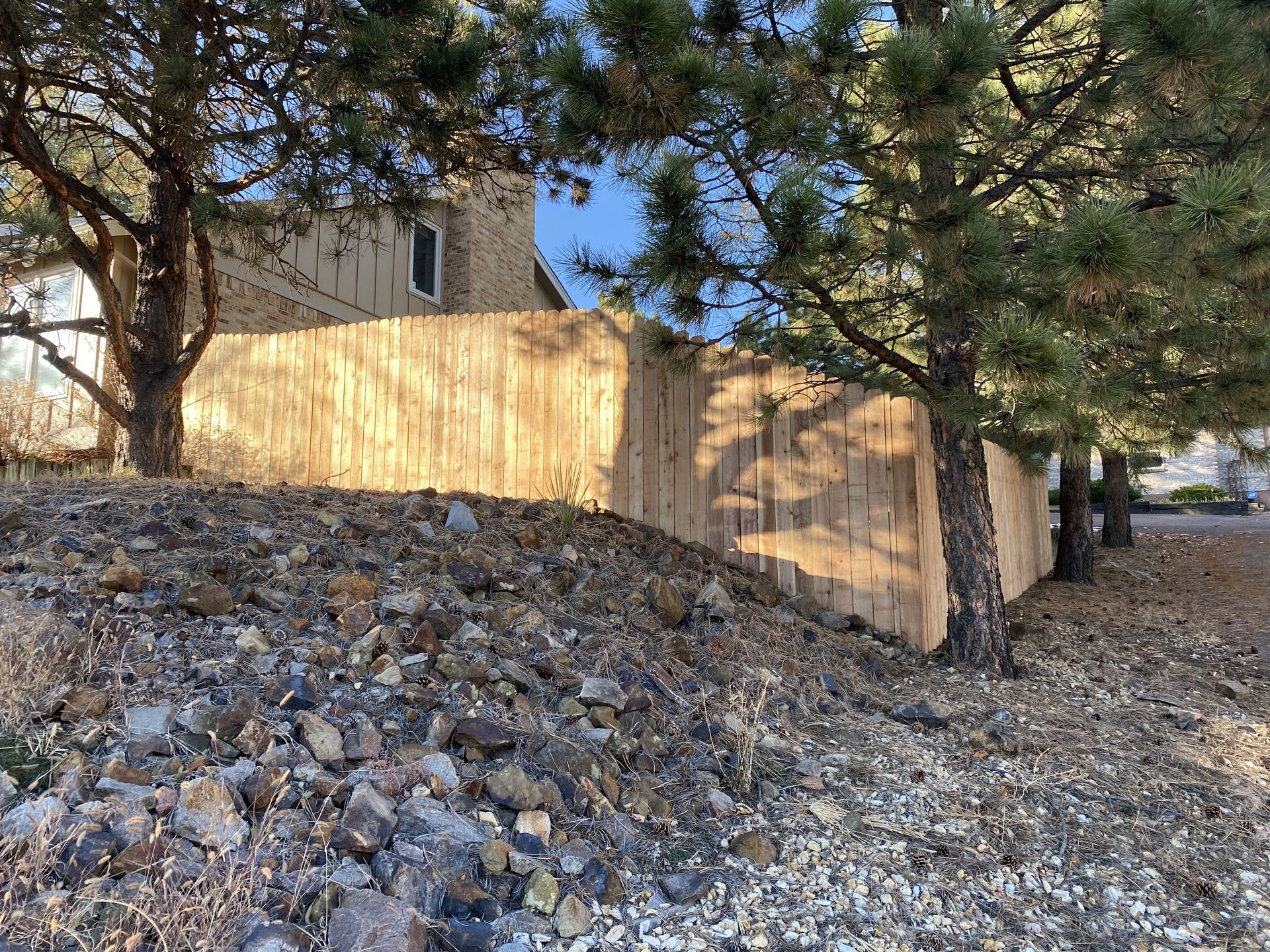 A wooden fence is surrounded by trees and rocks in front of a house.