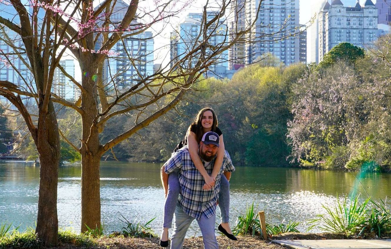 A man is carrying a woman on his back in front of a lake.