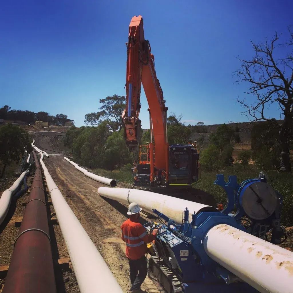 A Man Wearing A Hard Hat Stands Next To A Large Orange Excavator — Blackpoly Pipelines in Orange, NSW