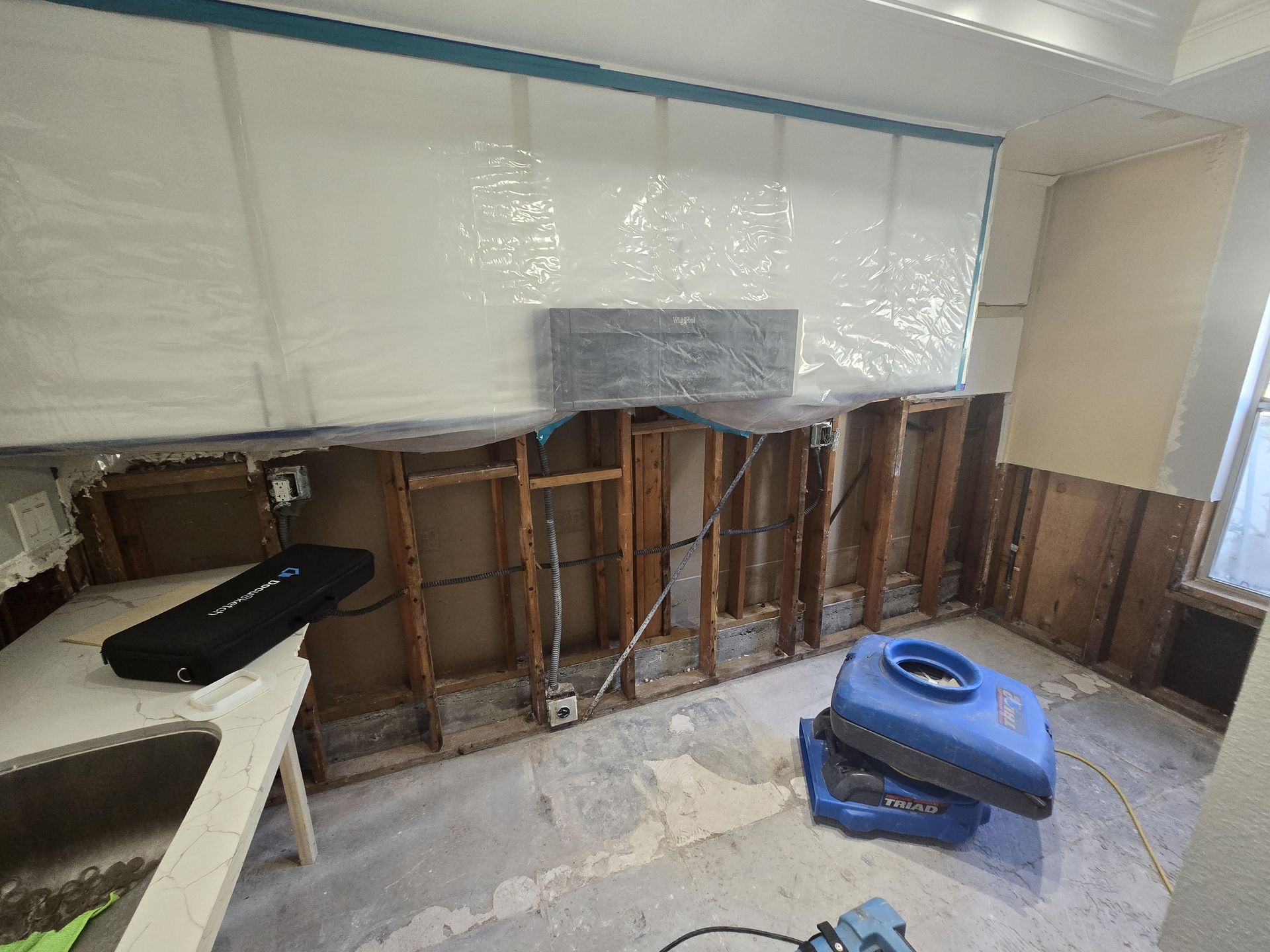 Kitchen undergoing water damage restoration, exposed framing, plastic sheeting, and drying equipment.