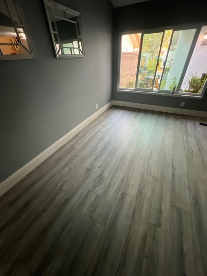 Empty room with grey walls, white baseboard, and wood-look flooring. Two mirrors hang on the wall, and a window is on the right.