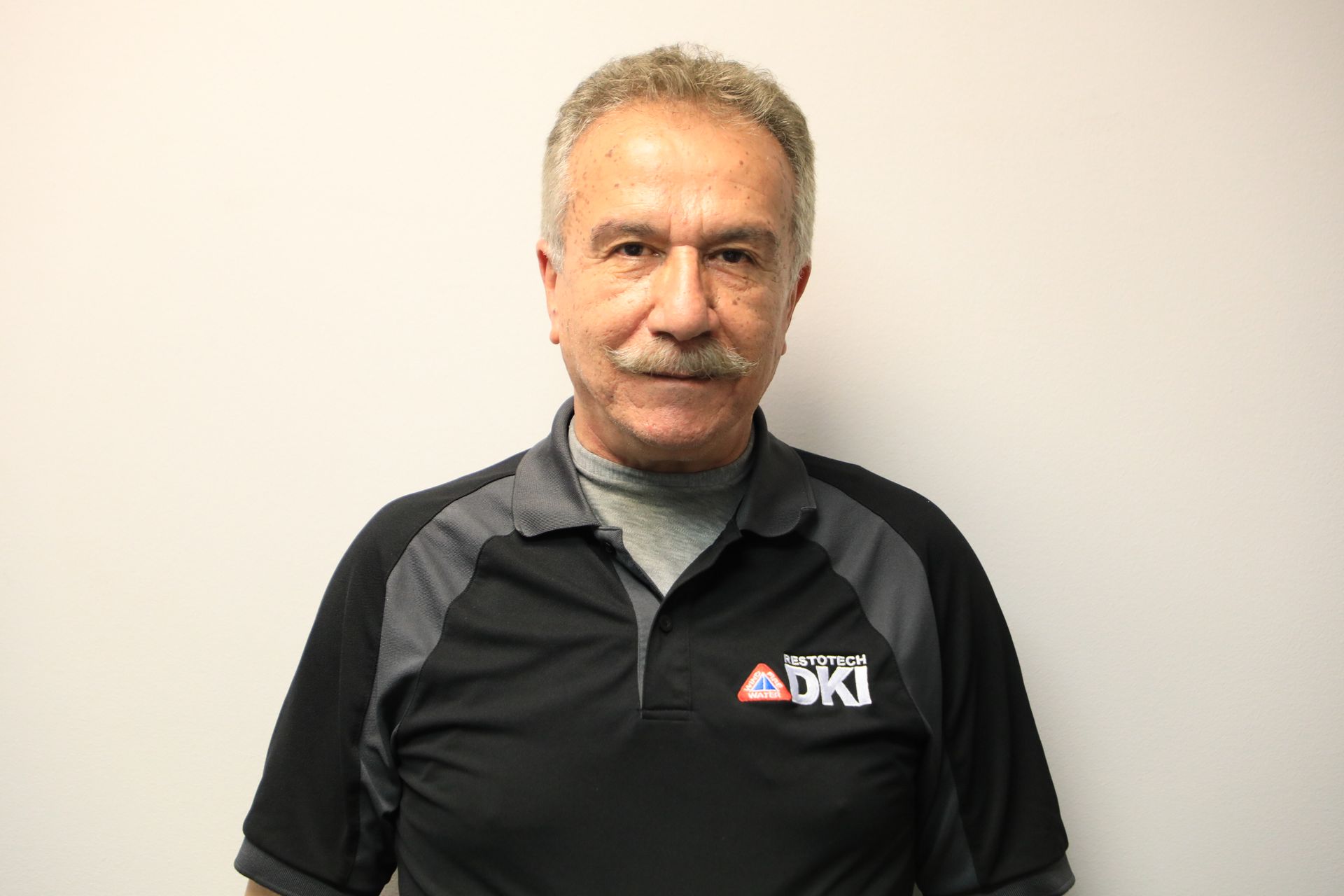 Man with graying hair and mustache wearing a black and gray polo shirt, standing in front of a white wall.