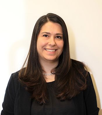 Woman with long dark hair smiles, wearing black blazer in front of a white wall.