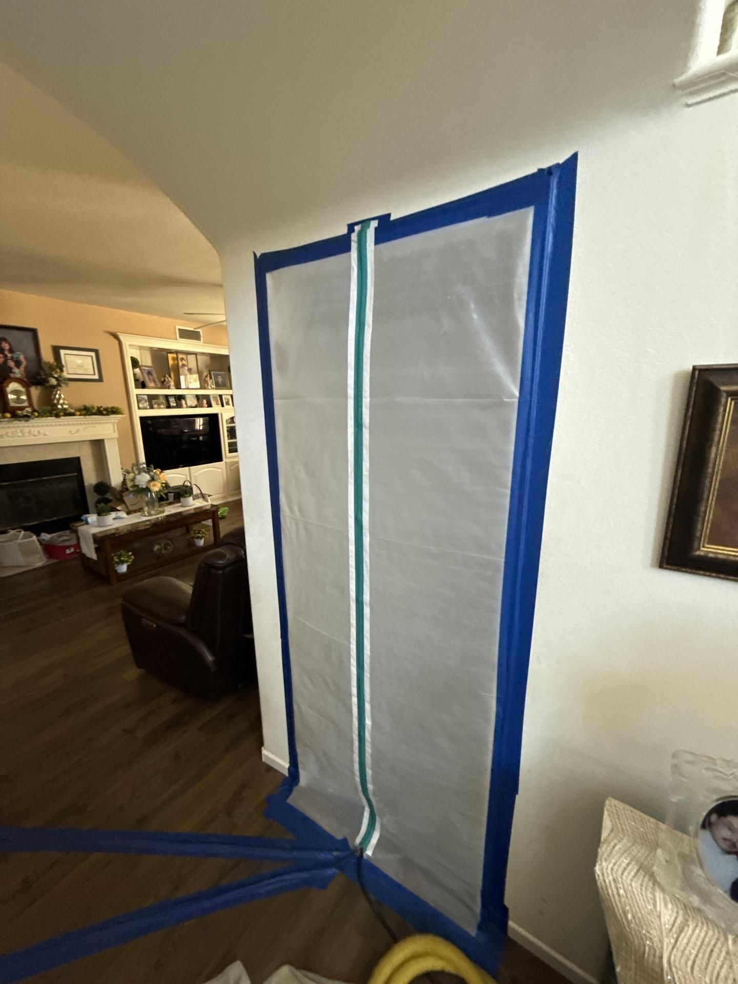 White plastic sheeting taped to a doorway, blocking the view into a living room. Blue tape borders the plastic.