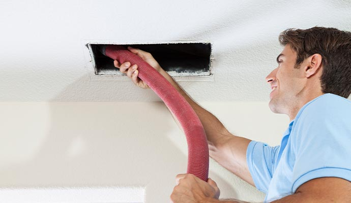 Man cleaning a vent in the ceiling with a hose, smiling. Pink hose, neutral setting.
