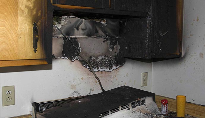 Charred kitchen cabinet and vent hood, likely from a fire. Damage visible on walls and surrounding area.