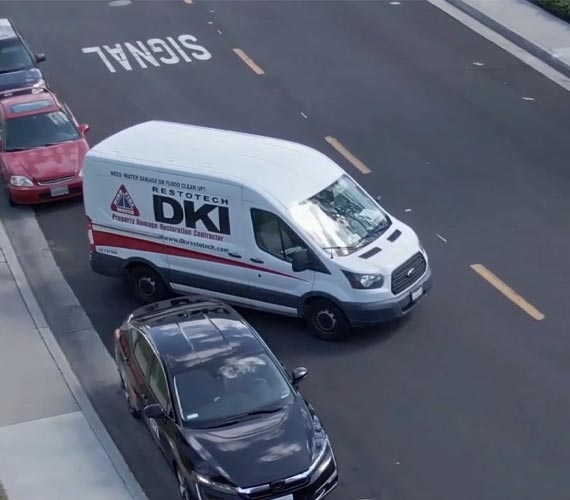 White van with DKI RESTORATION logo driving on a road next to parked cars, road markings.