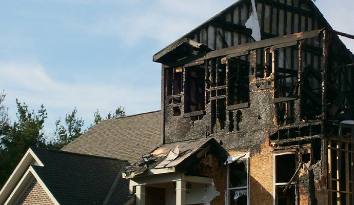House destroyed by fire, charred wooden frame and roof visible, daytime.