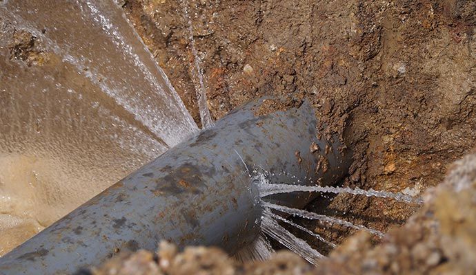 A broken gray pipe spurting water into a brown dirt trench.