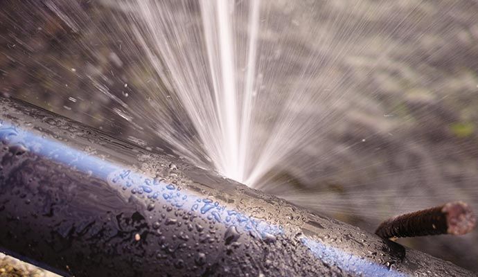 Water spraying forcefully from a punctured black pipe in an outdoor setting.