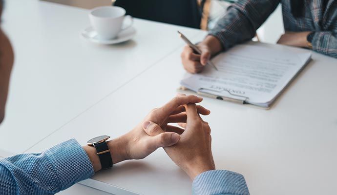 Person at a table with clasped hands, being interviewed by someone writing on a document.