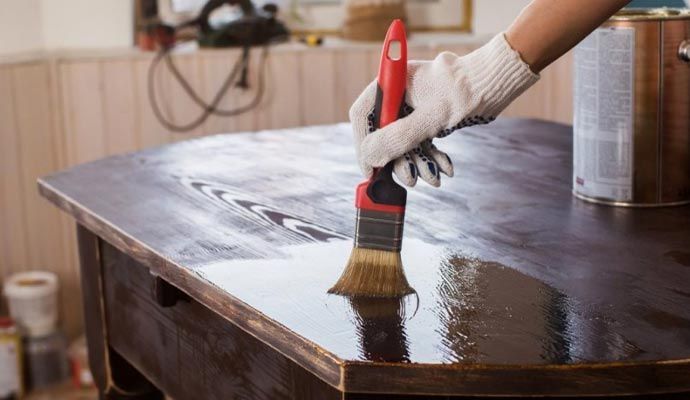 A person wearing gloves varnishes a dark brown wooden table with a paintbrush.