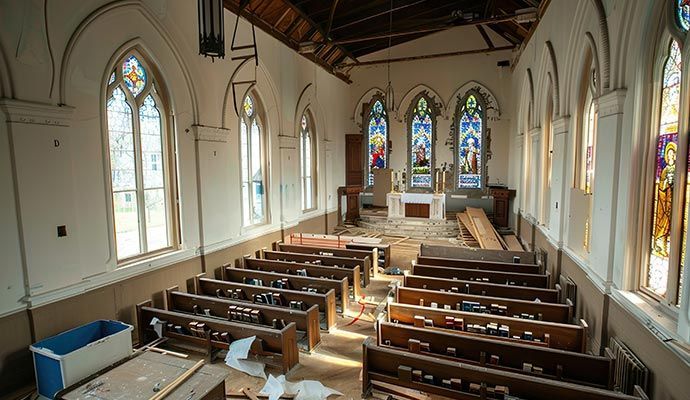 Interior of a dilapidated church with stained glass windows, rows of pews, and damaged ceiling.