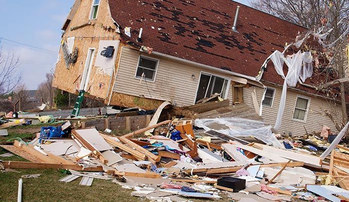 House severely damaged, debris scattered on lawn. Roof and walls torn away.