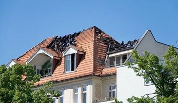 Burnt roof of a building with red tiles and open windows, against a blue sky.