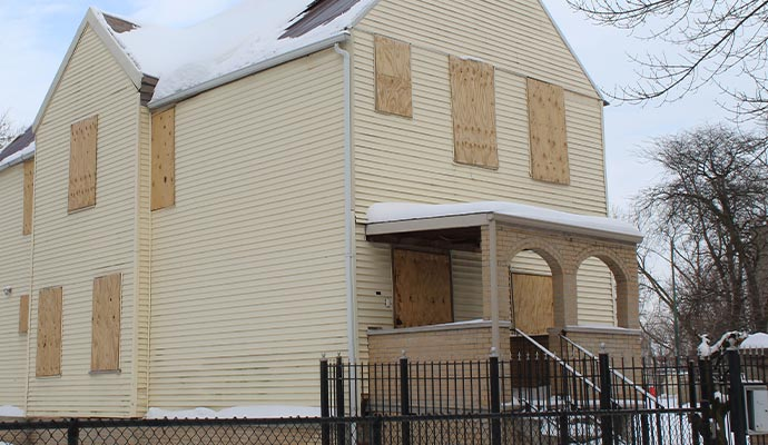 Two-story house with boarded-up windows, pale yellow siding, and a snow-covered porch, behind a black fence.