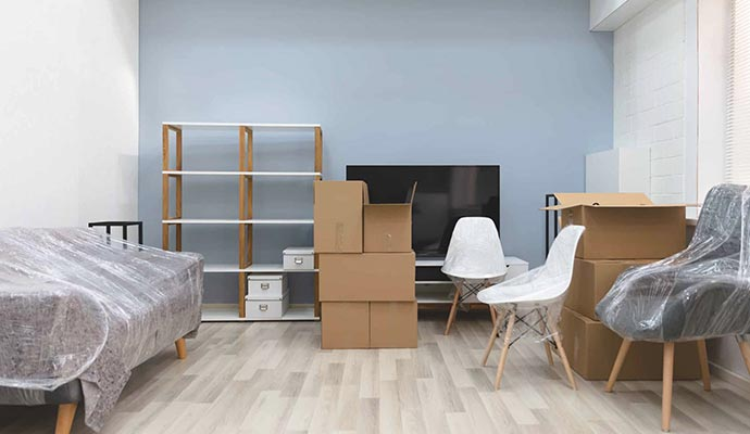 Living room with moving boxes, furniture wrapped in plastic, and a shelving unit against a blue wall.