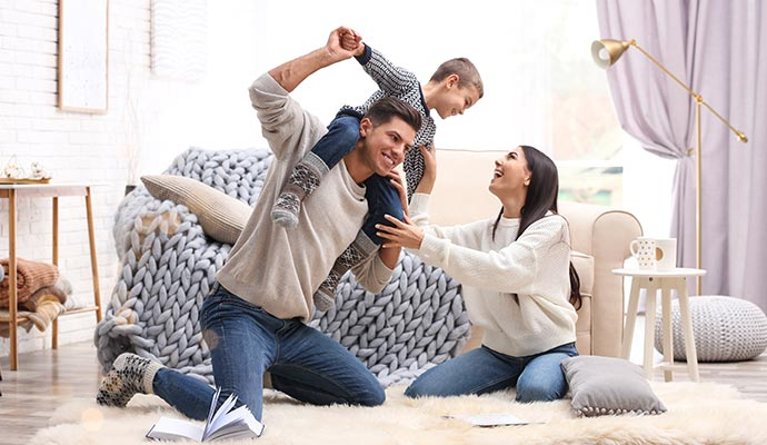 Man with child on shoulders, woman looks on smiling, in cozy living room.