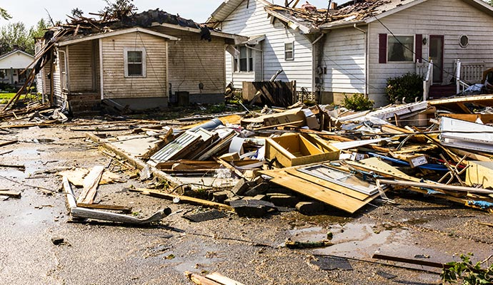 Houses with severe roof damage and debris-filled street, likely tornado aftermath.
