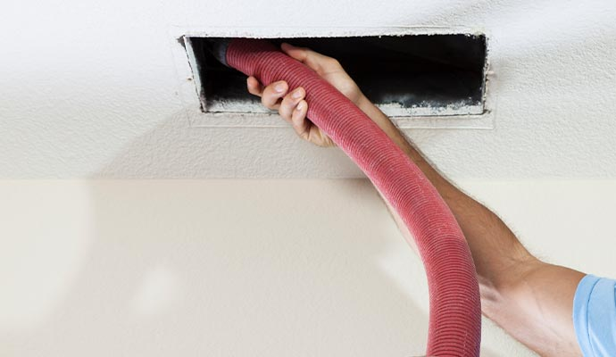 Person vacuuming an air vent in a ceiling with a red hose.