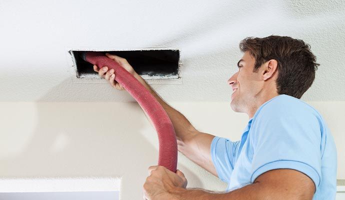 Man cleaning a ceiling vent with a long, red hose.