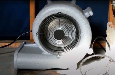 Large, silver industrial fan with a protective grill, on a reflective surface.