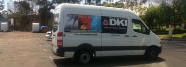 White van with DKI logo parked on a paved lot. Trees and buildings in the background.