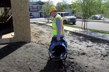 Construction worker in safety vest and hard hat carries a blue ventilation fan on a construction site.