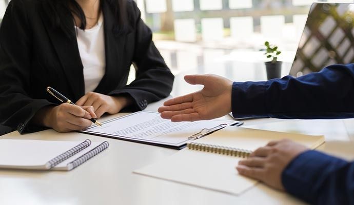 Person signing document across a desk, another person's hand extended toward them.
