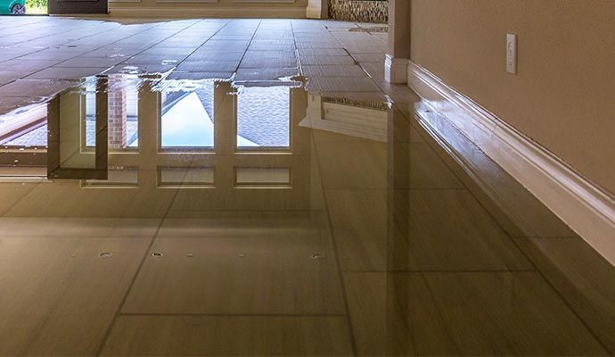 Flooded tile floor reflecting a window's white trim and a brown frame, near a tan wall with white baseboards.