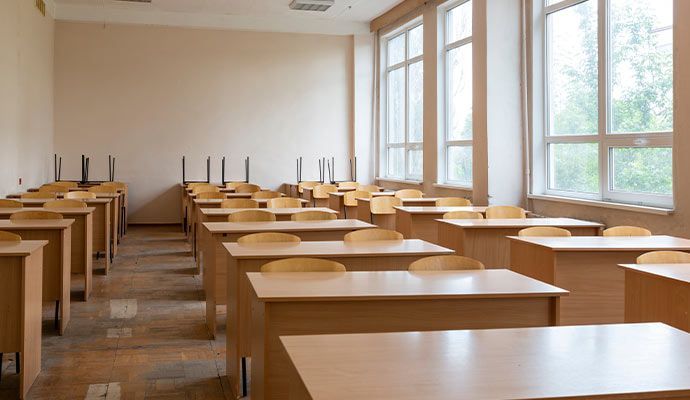 Empty classroom with rows of desks and chairs; large windows on the right.