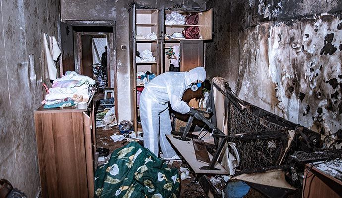 Person in white suit examines debris in a fire-damaged room with charred walls and furniture.