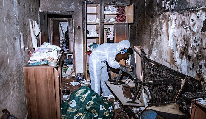 Person in protective suit examines fire-damaged room, charred furniture, debris, and burnt walls.
