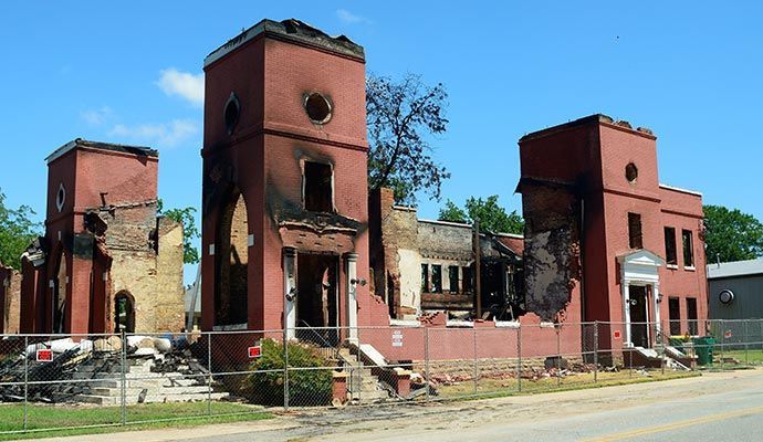 Fire-damaged red brick building with three towers; exterior destruction evident, fence in front.