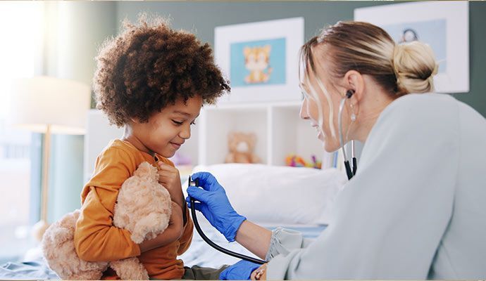 A young child with a teddy bear is being examined by a healthcare provider with a stethoscope.