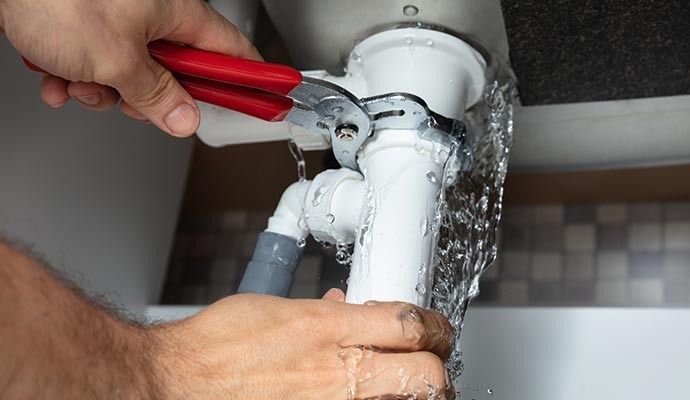 Plumber using a wrench on a leaky white sink drain pipe, water spraying out.