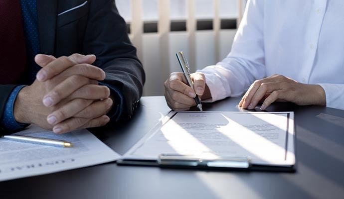 Person signing a document on a clipboard, another person's folded hands visible. Sunlight streams in.