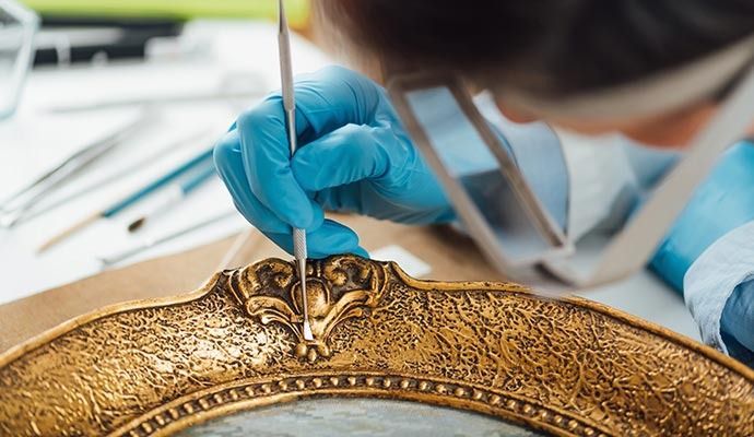 Person in blue gloves restoring a gold ornate picture frame with a small tool.