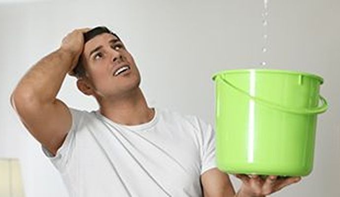 Man holding green bucket, looking up at a water leak in a white-walled room, concerned expression.